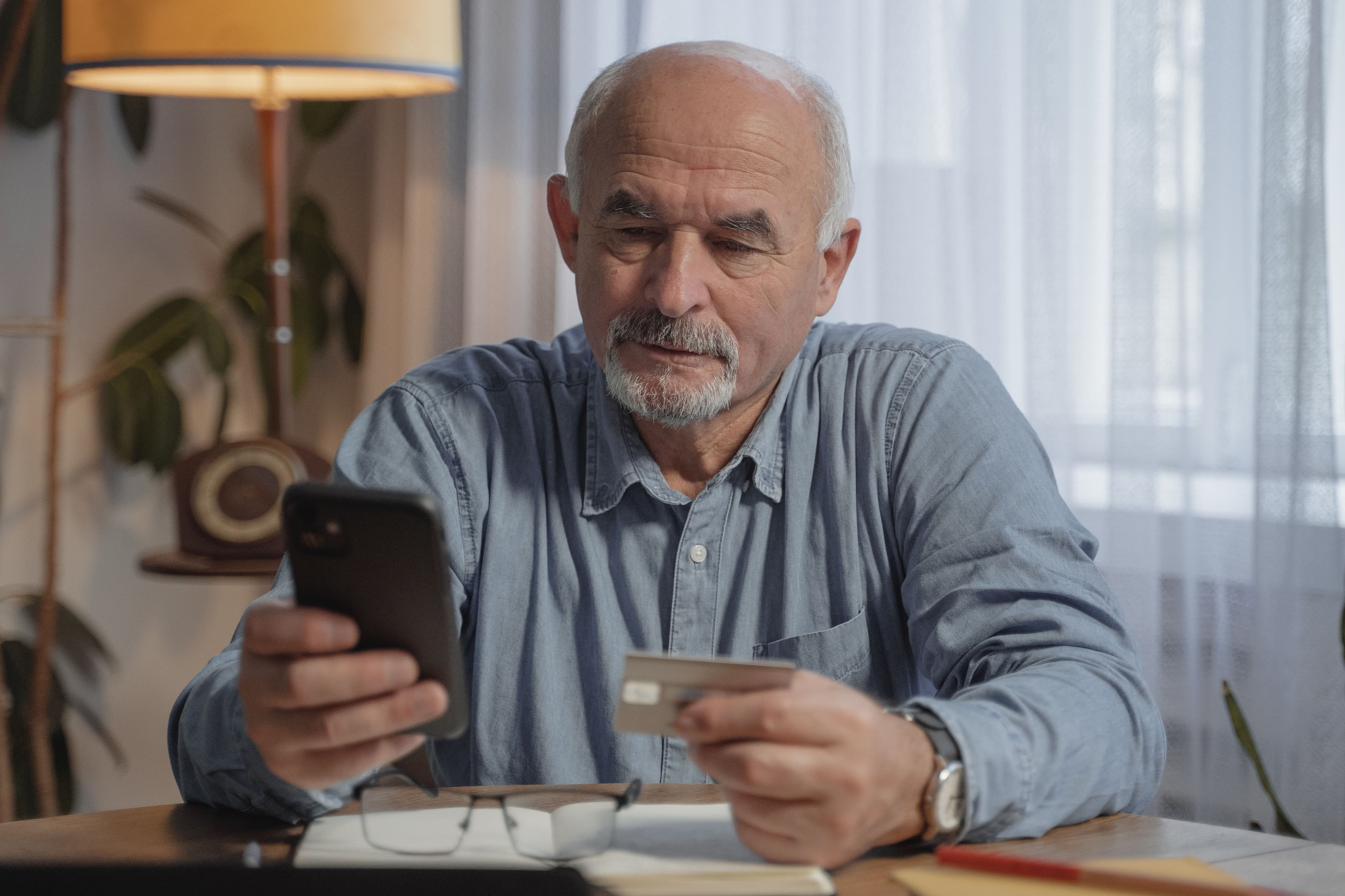 Older man using a smartphone while holding a payment card at home.